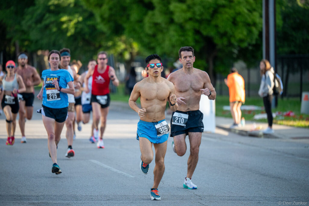 Group of runners competing in the Bellaire Trolley Run 5K road race on a tree-lined street in Bellaire, TX.