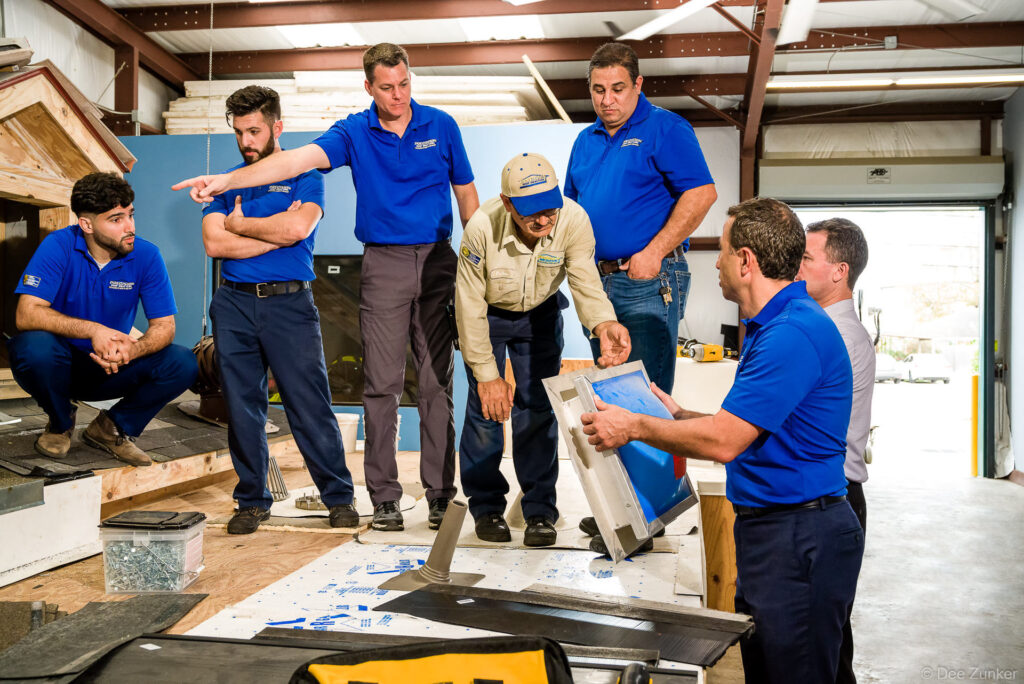 Roofing company branding photo showing six technicians in blue polo shirts reviewing materials and equipment in a Southwest Houston workshop