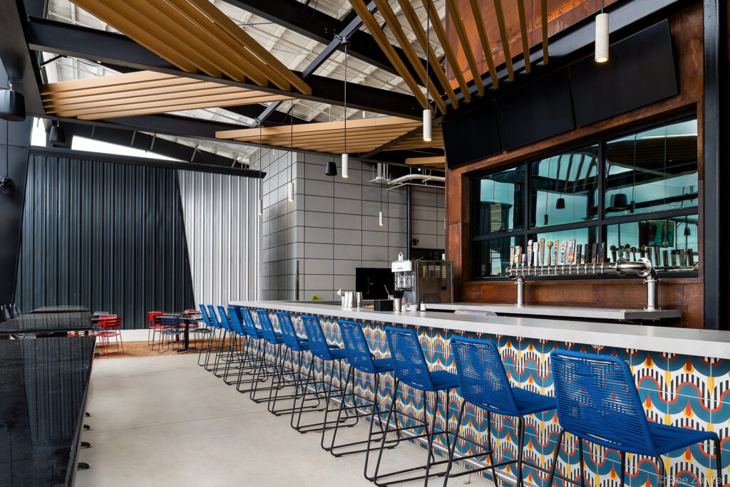 Modern brewery taproom bar with blue rope barstools, colorful geometric tile facade, draft beer taps, and wood slat ceiling in Katy TX