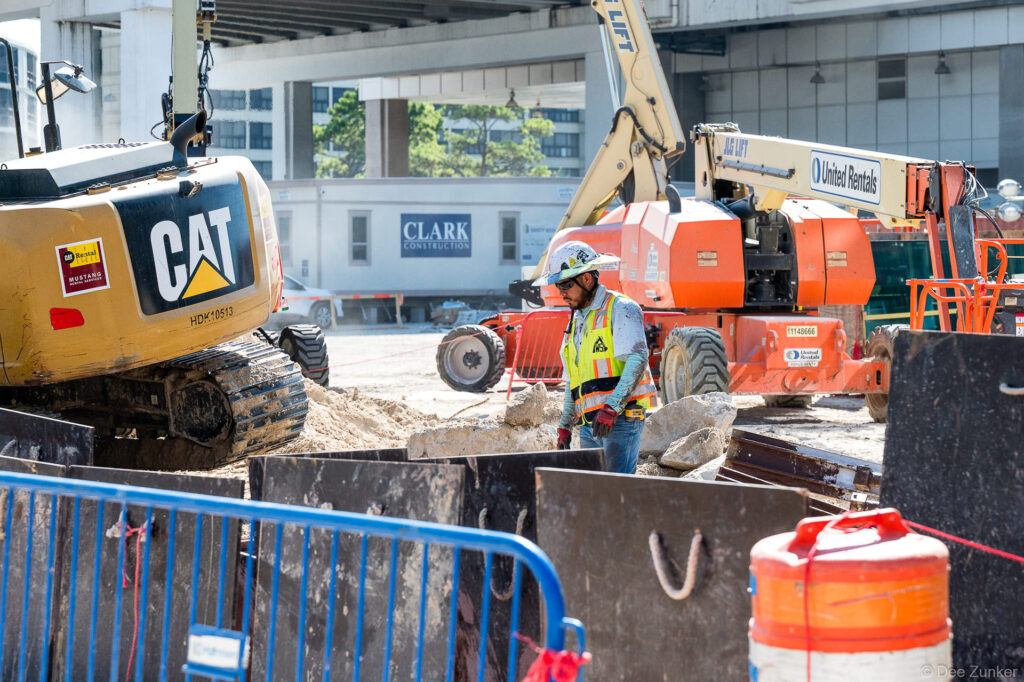 Construction worker in safety vest and hard hat stands beside a CAT excavator at the Clark Construction IAH B Terminal jobsite in Houston.