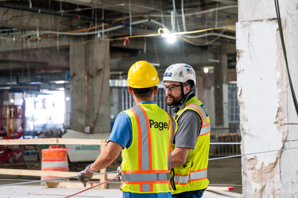 Two construction workers in high-visibility vests confer on the active jobsite interior of IAH Terminal B during structural progress work in Houston.