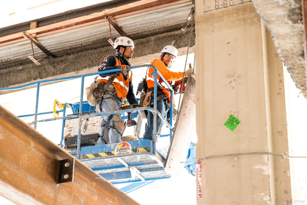 Two construction workers in orange high-vis gear inspect a concrete column from a Sunstate scissor lift at IAH Terminal B, Houston.