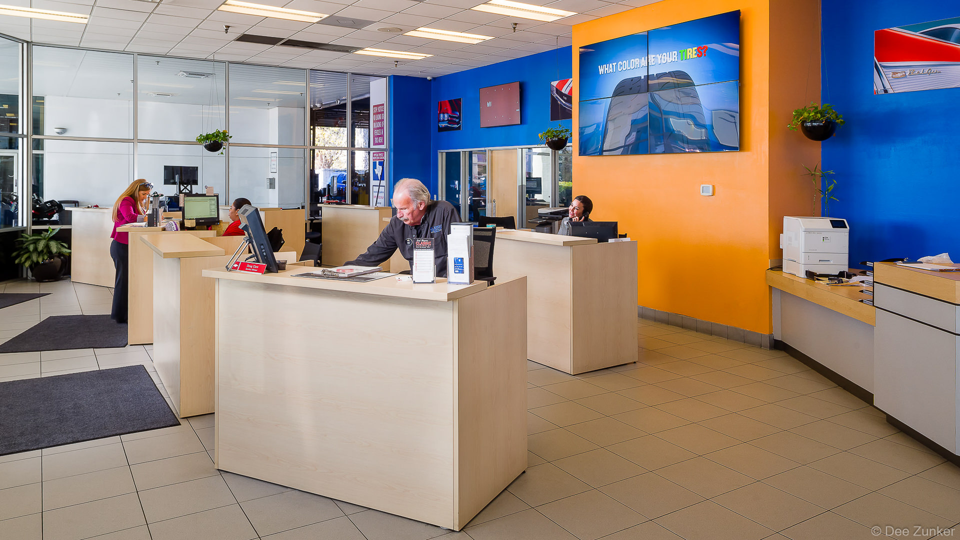 Bright dealership service counter interior with staff at workstations, blue and orange walls, and automotive displays in Houston, TX.