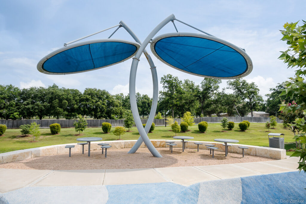 Architectural shade canopy with blue oval panels over picnic tables at Clements Crossing park, Houston TX, exterior landscape photography.