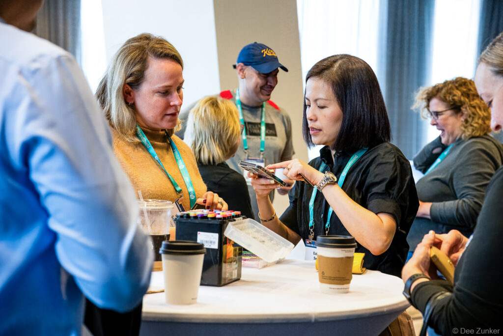 Conference attendees in teal lanyards gather around a table with markers and coffee cups during an interactive networking session.