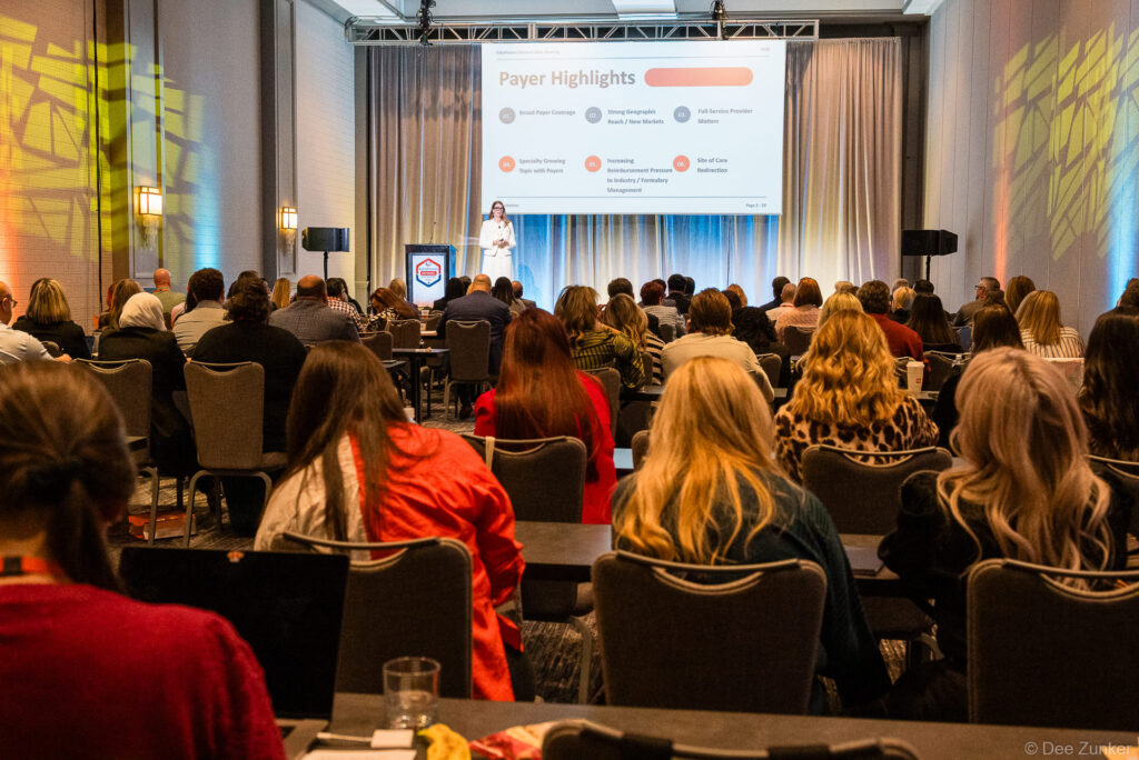 Speaker presents Payer Highlights slide to a full ballroom audience at a Houston hotel conference event, photographed by Dee Zunker Photography.