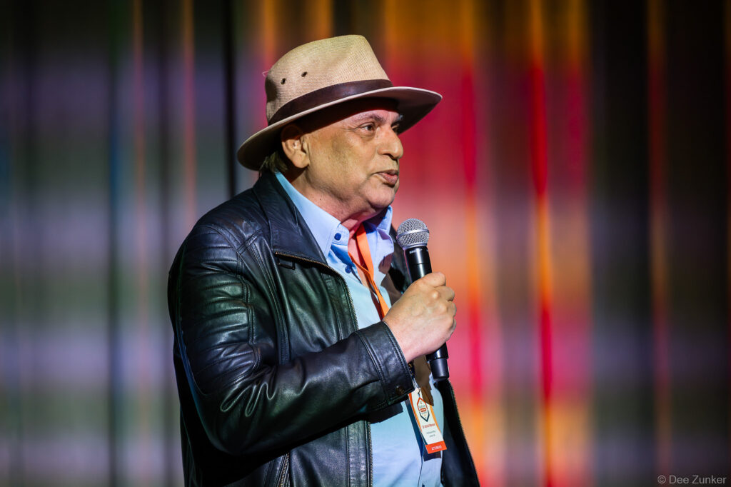 Male conference speaker in a fedora hat and leather jacket holds a microphone while addressing attendees at a Houston corporate conference event.