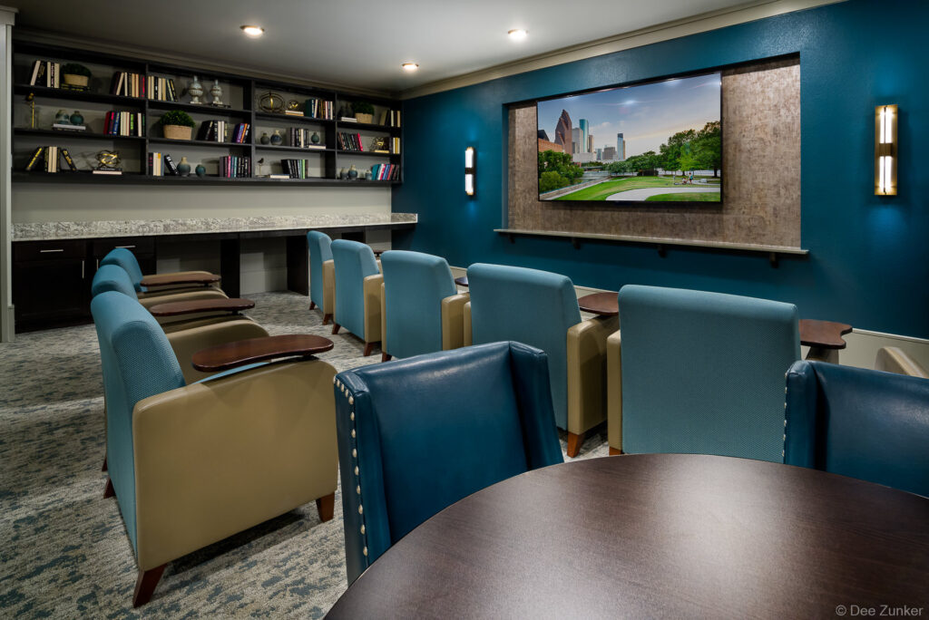 Modern senior apartment theater room with teal accent wall, large flat-screen TV, upholstered lounge chairs, and built-in bookshelves in Conroe TX