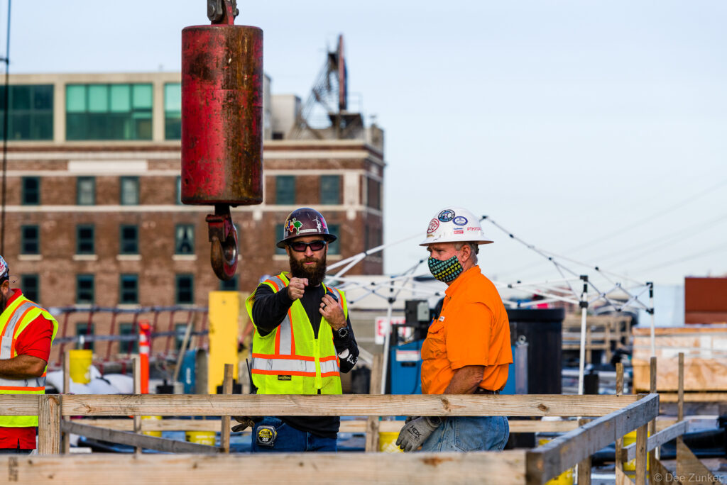 Two construction workers in hard hats and safety vests confer on an active commercial jobsite in Houston, with a crane hook overhead.