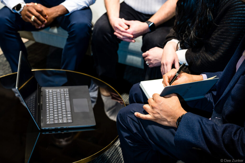 Business professionals seated in a circle during a corporate meeting, one taking notes in a notebook, laptop open on a gold-rimmed table.