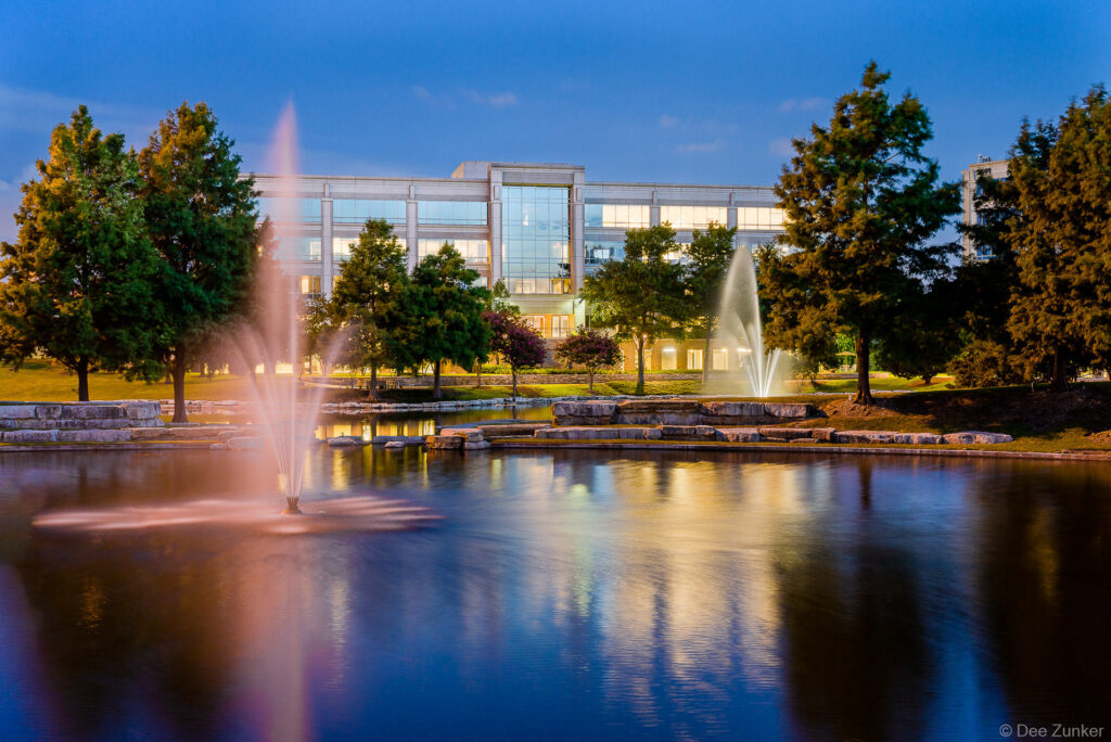 Corporate campus pond with illuminated fountains reflecting glass office building at blue hour, Houston commercial exterior photography