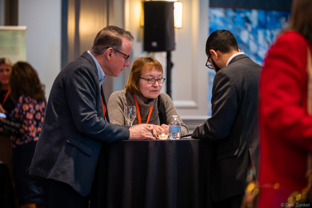 Three conference attendees in business attire engage in conversation at a standing table during a corporate networking event.