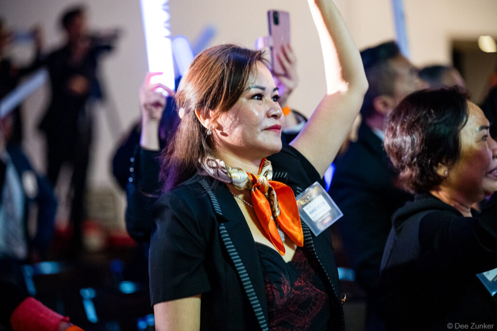 Woman in black blazer and orange scarf raises glowing foam stick at a Houston corporate event gala, crowd visible behind her.