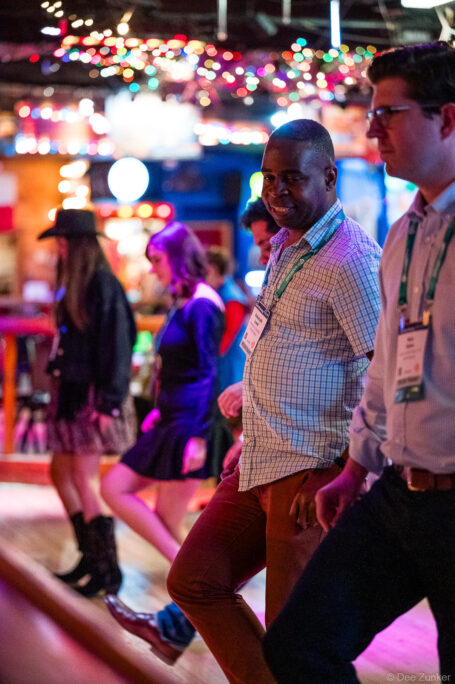 Conference attendees with lanyards and name badges walk through a colorfully lit Houston corporate event venue at night.