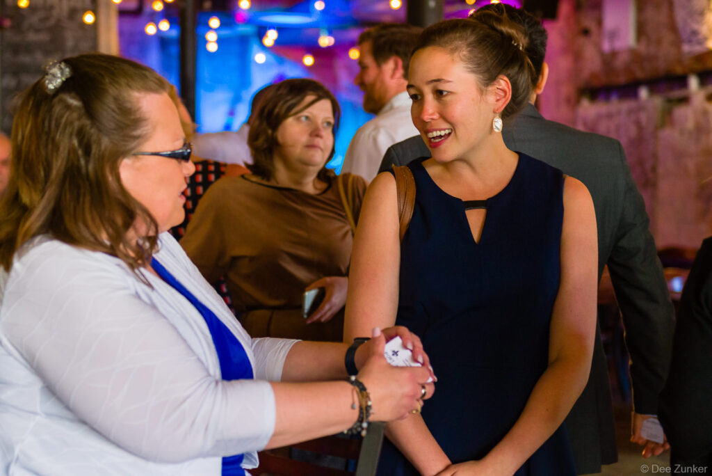 Two women shake hands and smile during a Houston corporate event, with other attendees visible in a warmly lit venue background.
