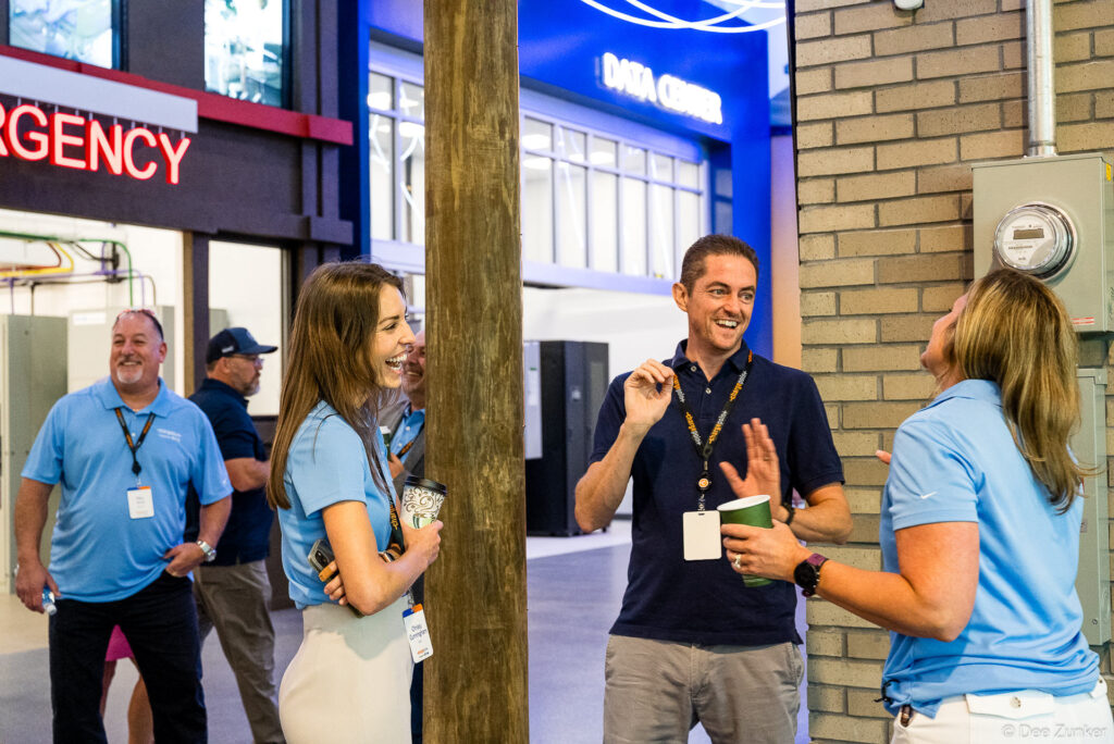 Professionals in light blue polo shirts laugh and chat near a illuminated data center exhibit at a Houston corporate event.