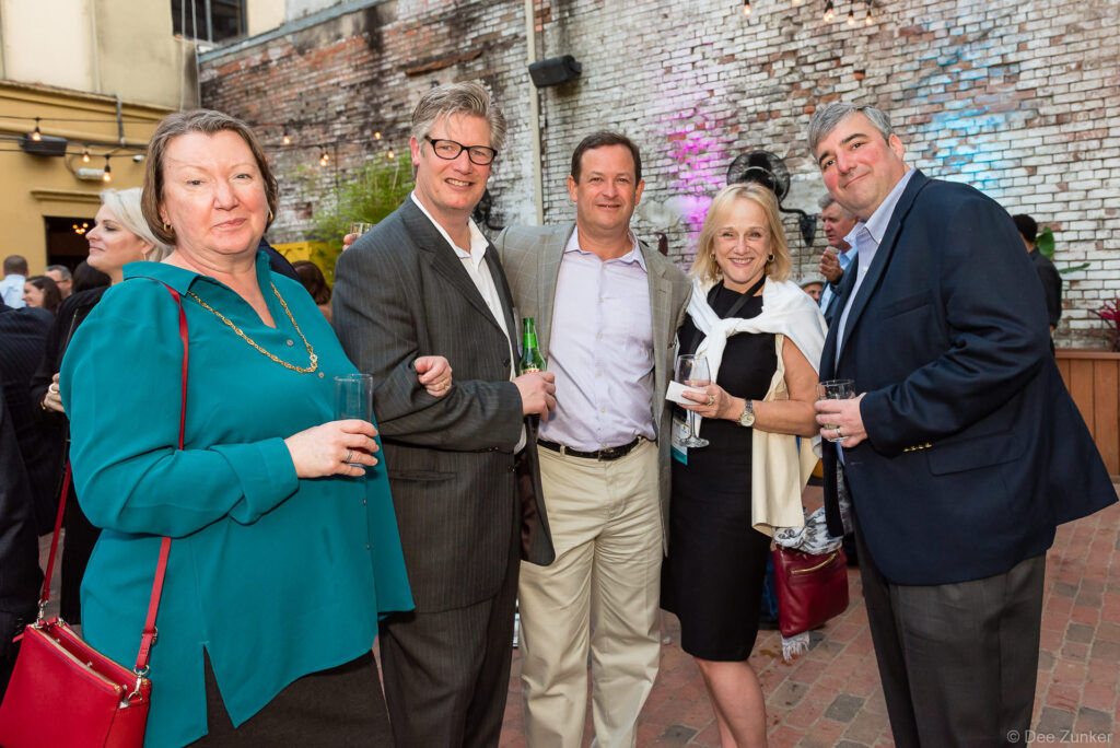 Five corporate event guests smile and hold drinks at an outdoor Houston networking reception with exposed brick walls.