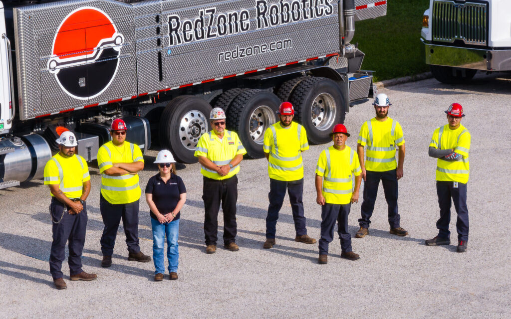 Eight industrial workers in hi-vis vests and hard hats pose before a branded robotics service truck on a gravel lot.