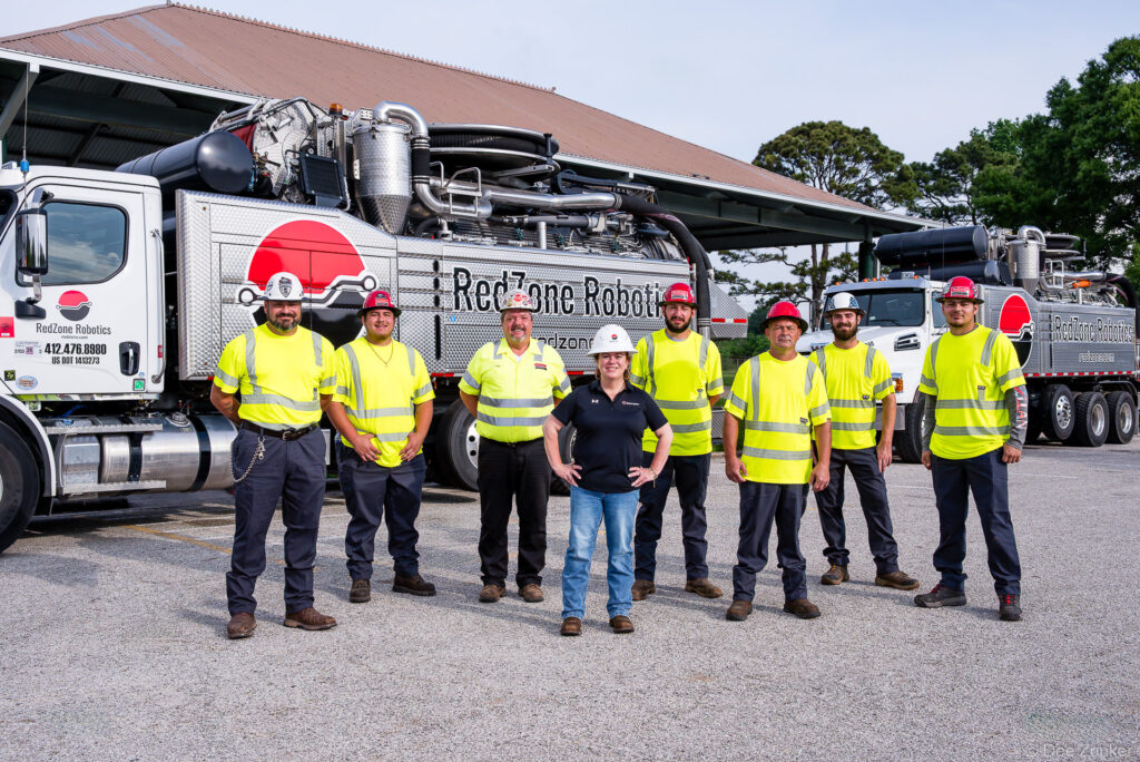 Eight corporate field team members in hi-vis safety gear and hard hats pose before branded industrial vacuum trucks outdoors.