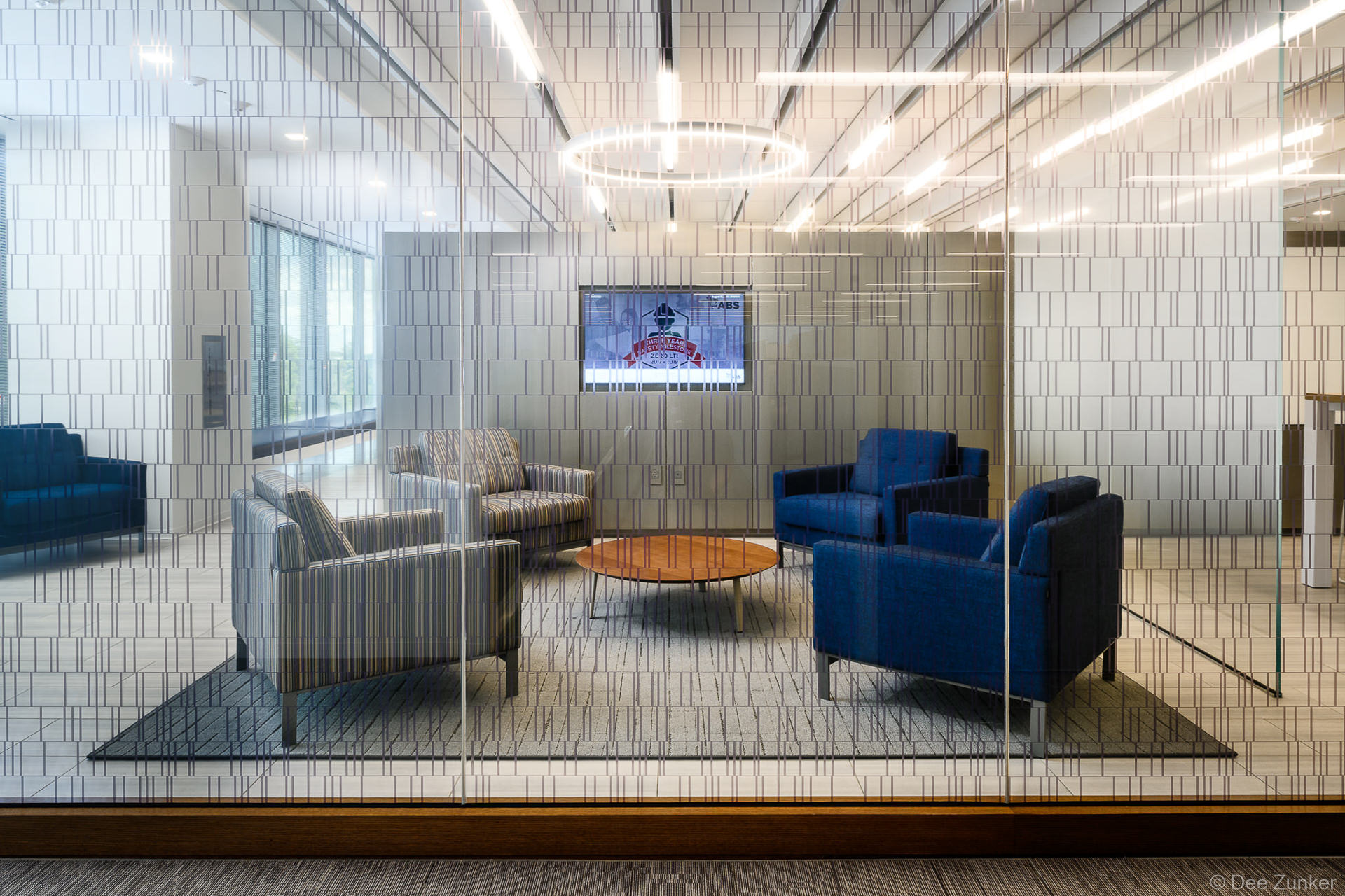 Glass-walled corporate lounge with blue and gray armchairs, round wood coffee table, and wall-mounted display screen