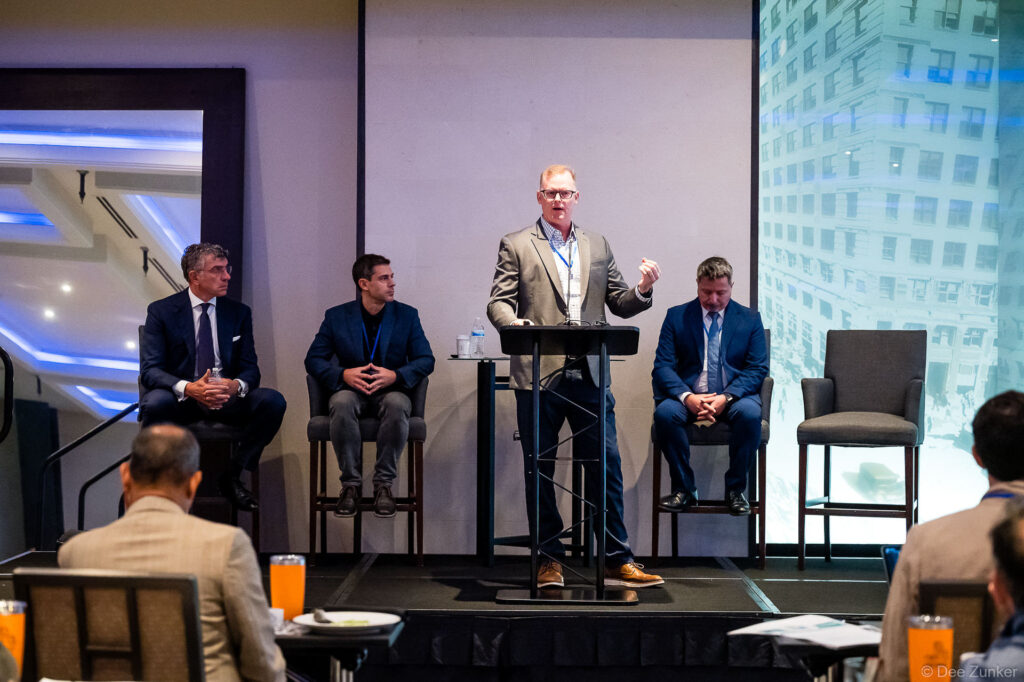 Speaker at podium addresses corporate conference panel with three seated panelists on stage, Houston professional event photography.