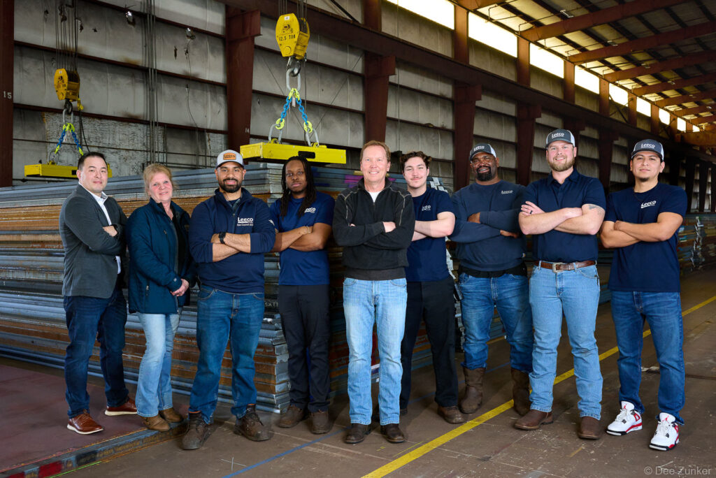 Nine corporate team members pose with arms crossed inside a large Houston steel distribution warehouse facility for a professional group portrait.