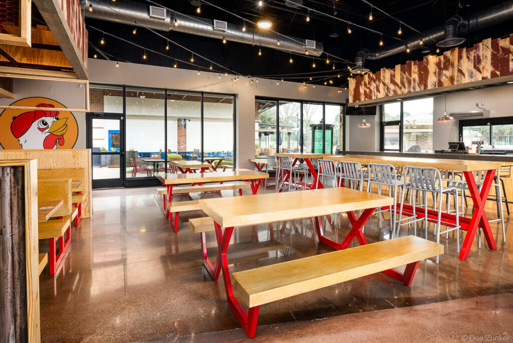 Bright restaurant interior with wood-top picnic tables, red metal legs, string lights, and rooster mural in Katy Texas.