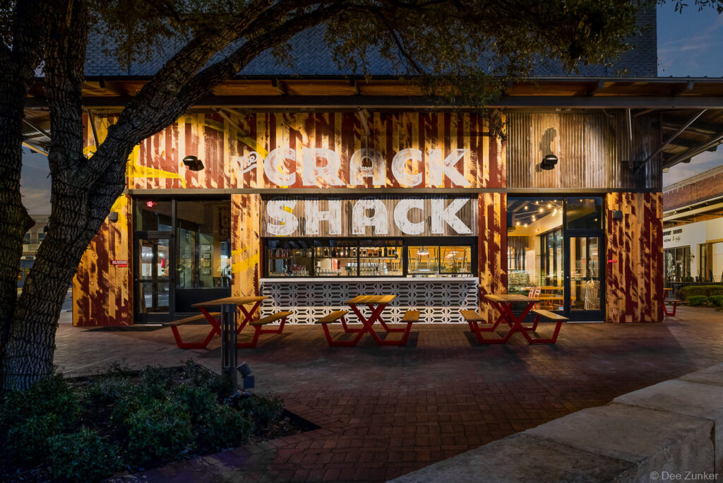 Crack Shack restaurant exterior at twilight featuring rustic corrugated metal facade bold signage and red picnic tables in Katy Texas