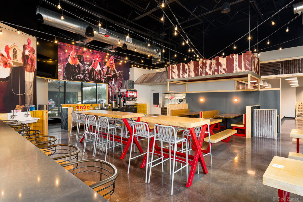 Casual fast-casual restaurant dining room with red picnic tables, metal bar stools, string lights, and bold chicken-themed wall murals in Katy TX