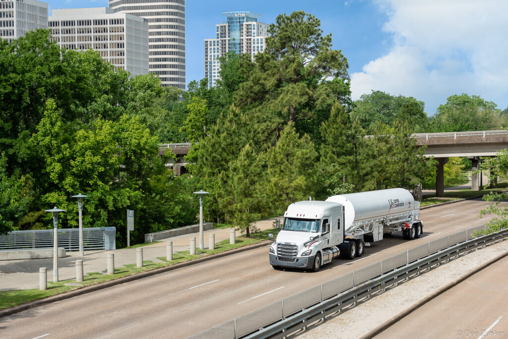 White cryogenic tanker truck traveling on a Houston urban highway surrounded by trees and city skyline, commercial transport photography.