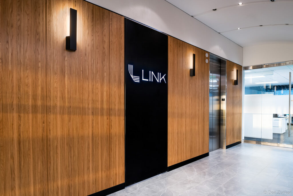 Modern corporate office lobby in Dallas featuring wood-paneled walls, illuminated wall sconces, elevator bank, and branded signage in a polished corridor.