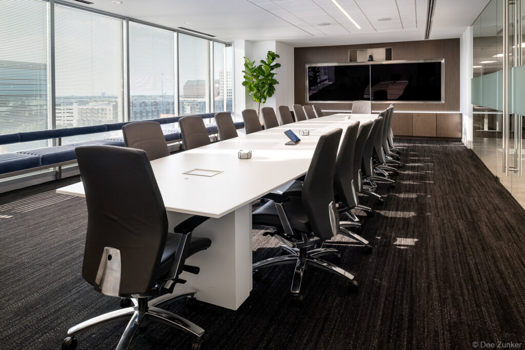 Executive conference room with long white table, leather chairs, large display screen, and floor-to-ceiling windows overlooking Dallas skyline