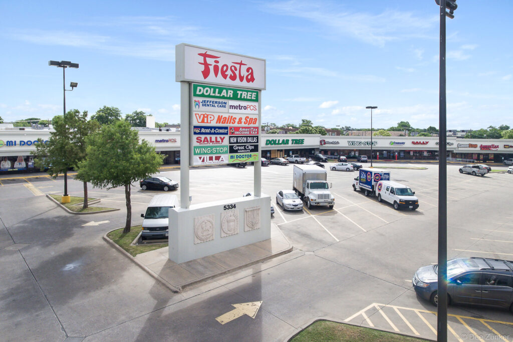 Aerial view of a Dallas TX strip mall parking lot with pylon sign listing multiple retail tenants on a sunny day.