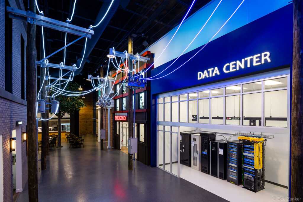 Indoor experience center exhibit featuring a data center display with server racks, power utility poles, and glowing cable lines in Houston.