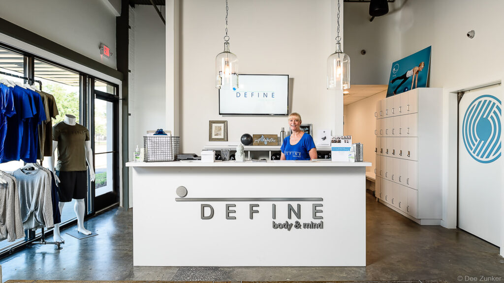 Modern fitness studio reception desk with smiling staff member, white lockers, retail area, and branded signage in Bellaire Houston