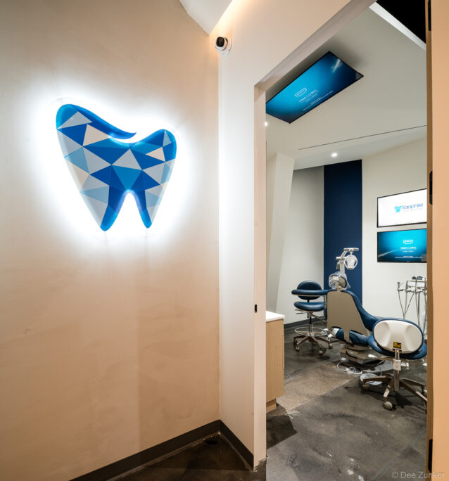 Illuminated geometric tooth logo wall art outside a modern dental treatment room with blue chair and ceiling monitor in Katy TX.