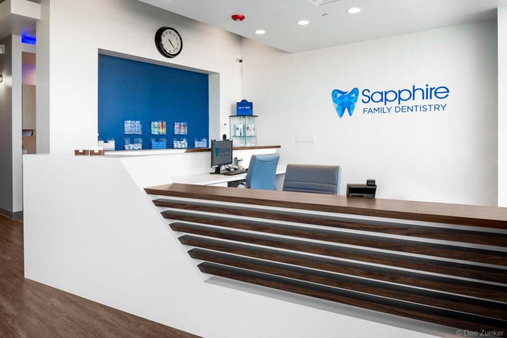 Modern dental office reception desk with wood accents, blue chairs, and branded wall signage photographed by Houston commercial photographer.