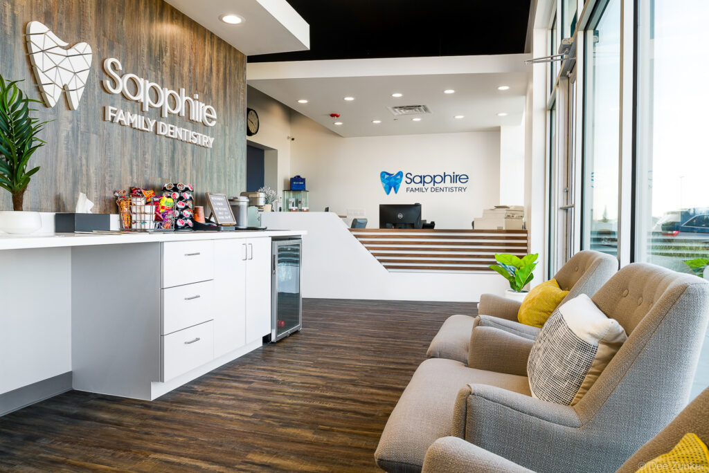 Modern dental office reception area with wood accent wall, white cabinetry, gray lounge chairs, and dual front desk areas in Houston, TX.