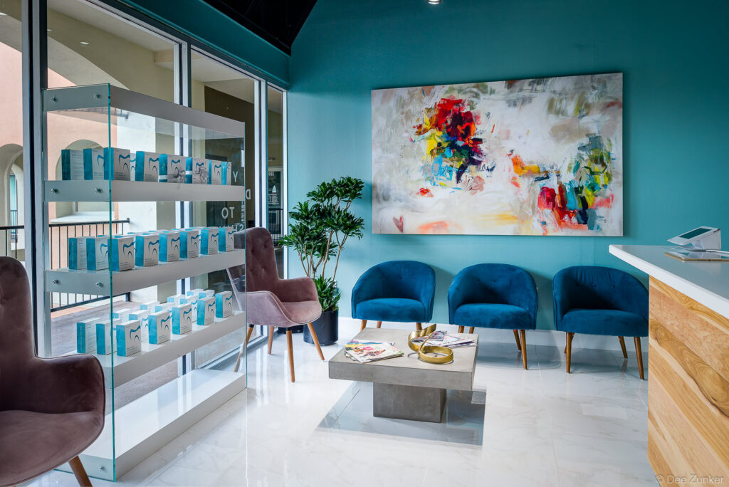 Modern dental office waiting room with teal walls, blue velvet chairs, abstract artwork, and product display shelving in Houston Galleria area