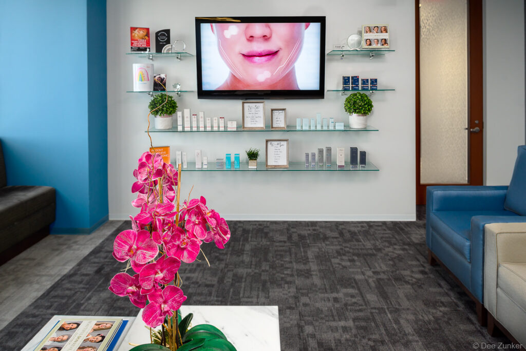 Modern dermatology clinic waiting room in Houston with product shelves, orchid centerpiece, and TV displaying facial treatment animation.