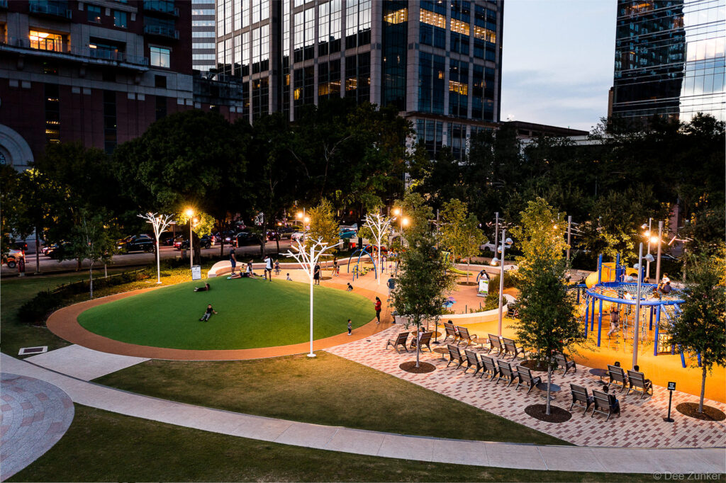 Aerial dusk view of Discovery Green park in Houston showing illuminated playground, lawn, and seating areas surrounded by downtown skyscrapers.