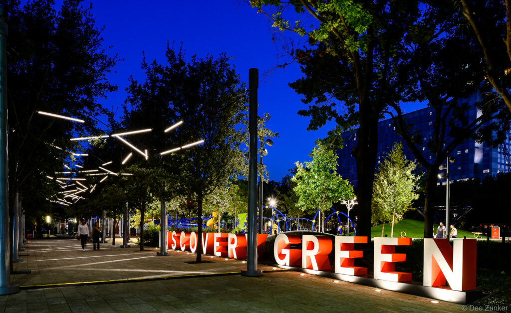 Large red Discovery Green sign illuminated at night with geometric LED light installations along a tree-lined promenade in Houston, TX.