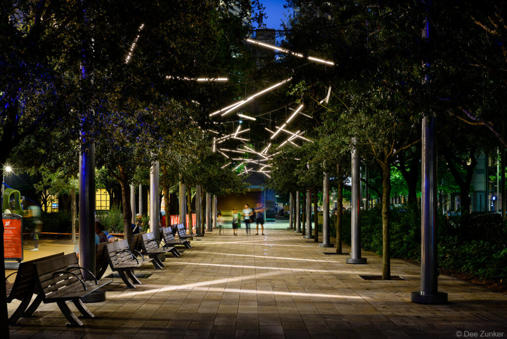 Discovery Green park walkway at night featuring suspended linear LED light installation among tree-lined promenade in downtown Houston