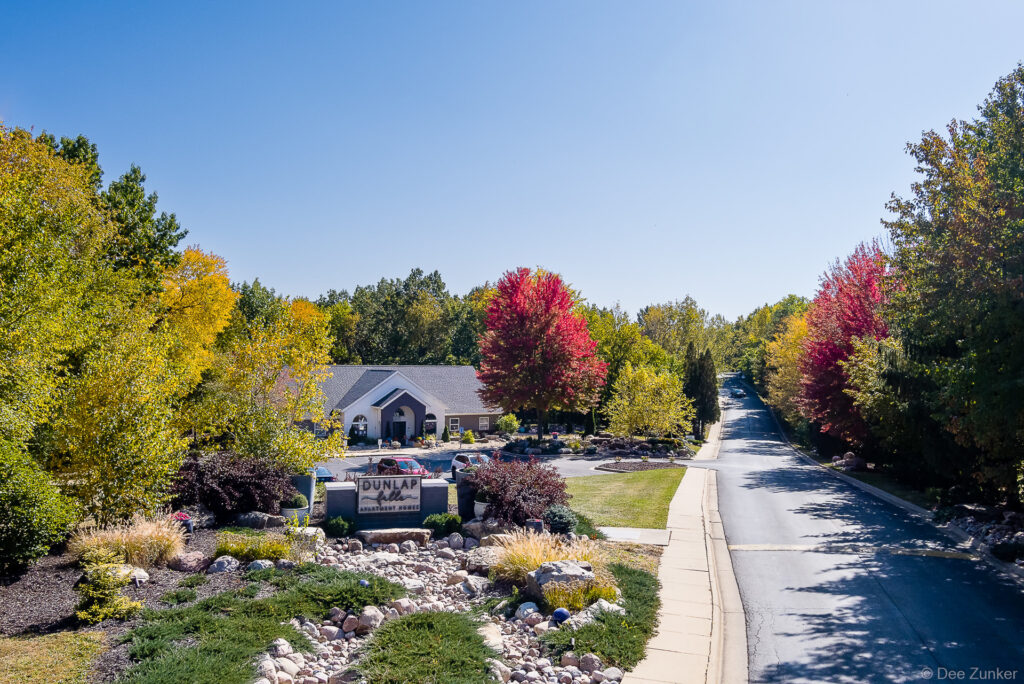 Aerial view of Dunlap Falls apartment community entrance in Illinois, featuring vibrant fall foliage, landscaped rock garden, and tree-lined road.