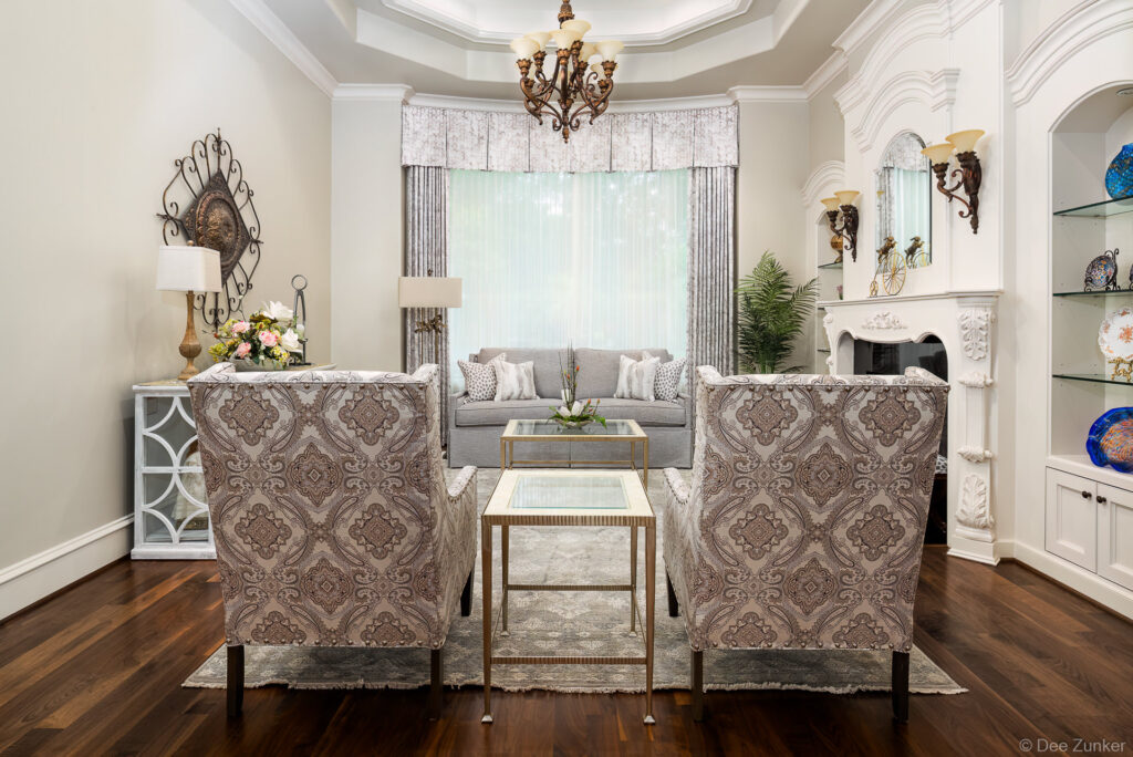 Formal living room with patterned wingback chairs, gray sofa, chandelier, ornate fireplace, and decorative accents in Houston home.