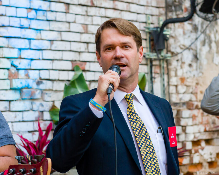 Male executive in navy suit and gold tie speaks into microphone at outdoor Houston corporate event against rustic brick wall backdrop.
