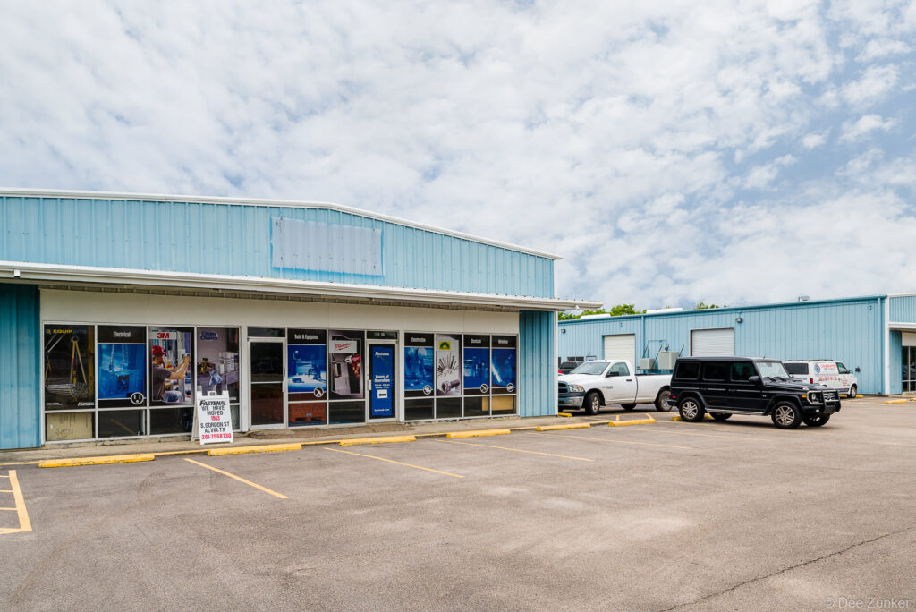Exterior of a light blue metal industrial building housing a fastener supply retailer on North Main Street, Houston TX, with storefront window displays and parked vehicles.
