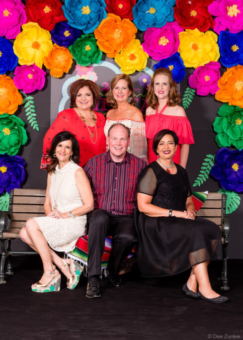 Six corporate event attendees pose on a bench in front of a colorful paper flower backdrop at Fiesta Flower Mound Houston area.