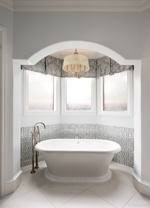 Elegant white freestanding soaking tub in an arched alcove with mosaic tile surround, chandelier, and bay windows in a Houston interior.