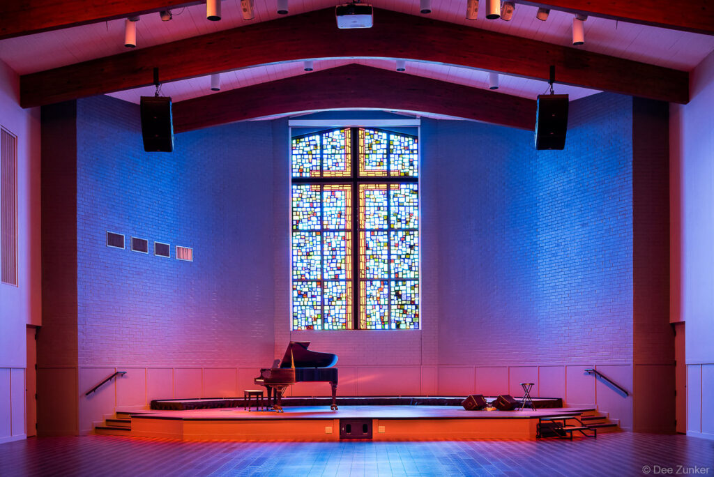 Church worship hall interior with grand piano on stage, colorful stained glass cross window, and dramatic blue and red LED lighting